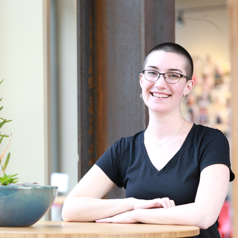 a pale woman in a black t-shirt with a shaved head and glasses smiling with her arms folded